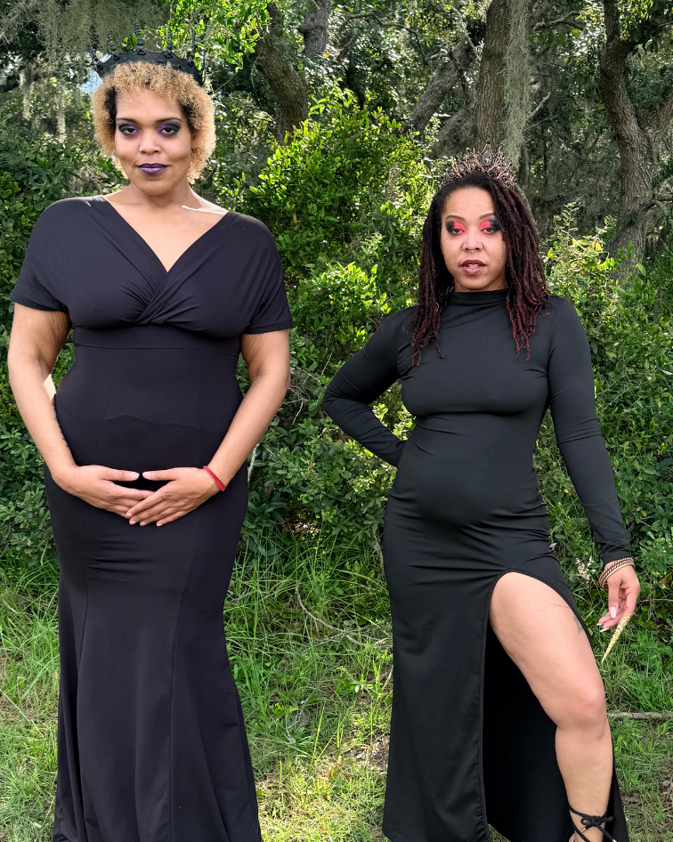 C.R.O.W.N. Jewels posing outdoors in matching black gothic-style dresses, standing on grassy ground with trees in the background. A gold monogram logo is visible in the bottom right corner.